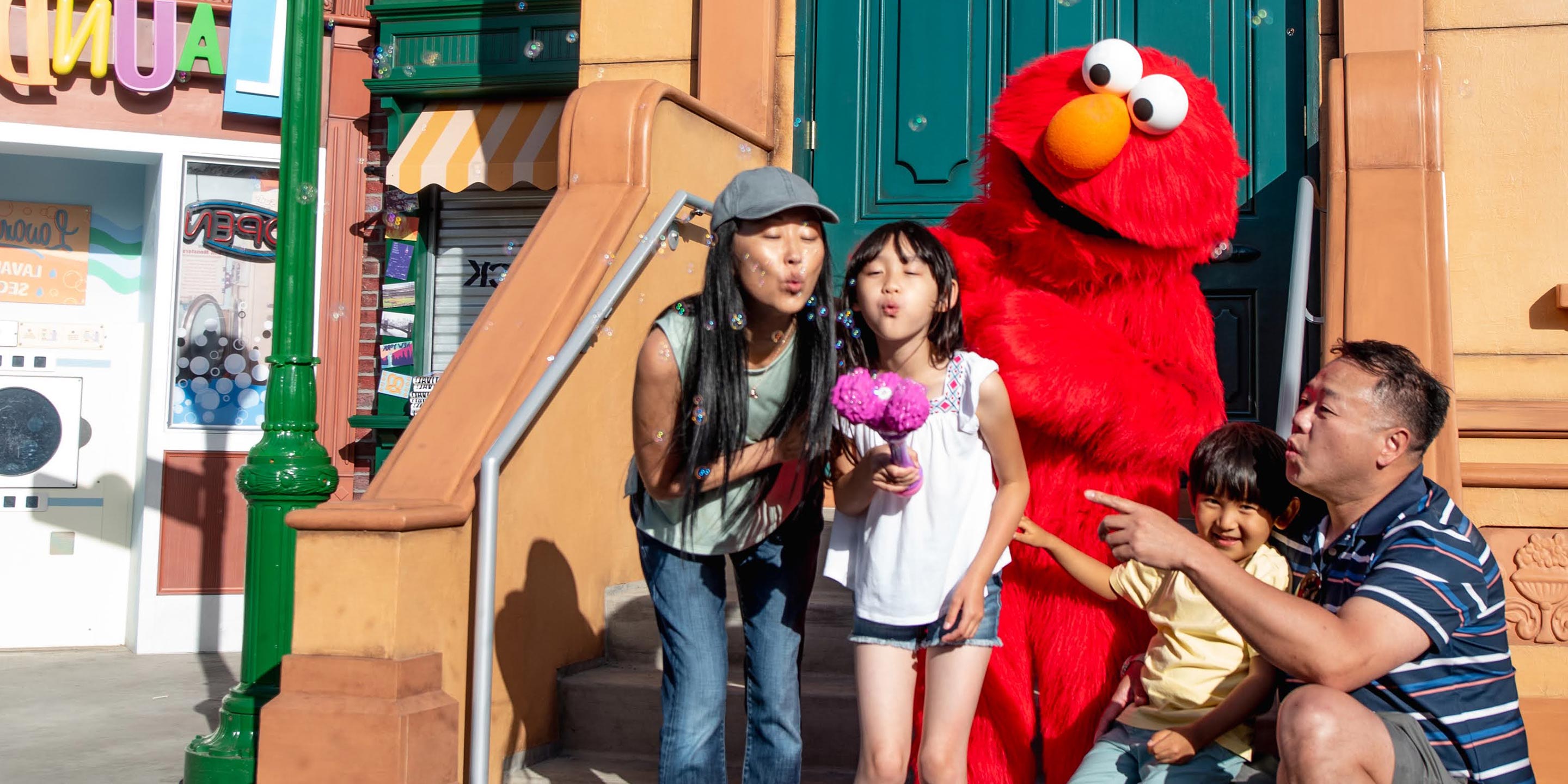 A family sits on the stairs, blowing bubbles into the air, with Elmo